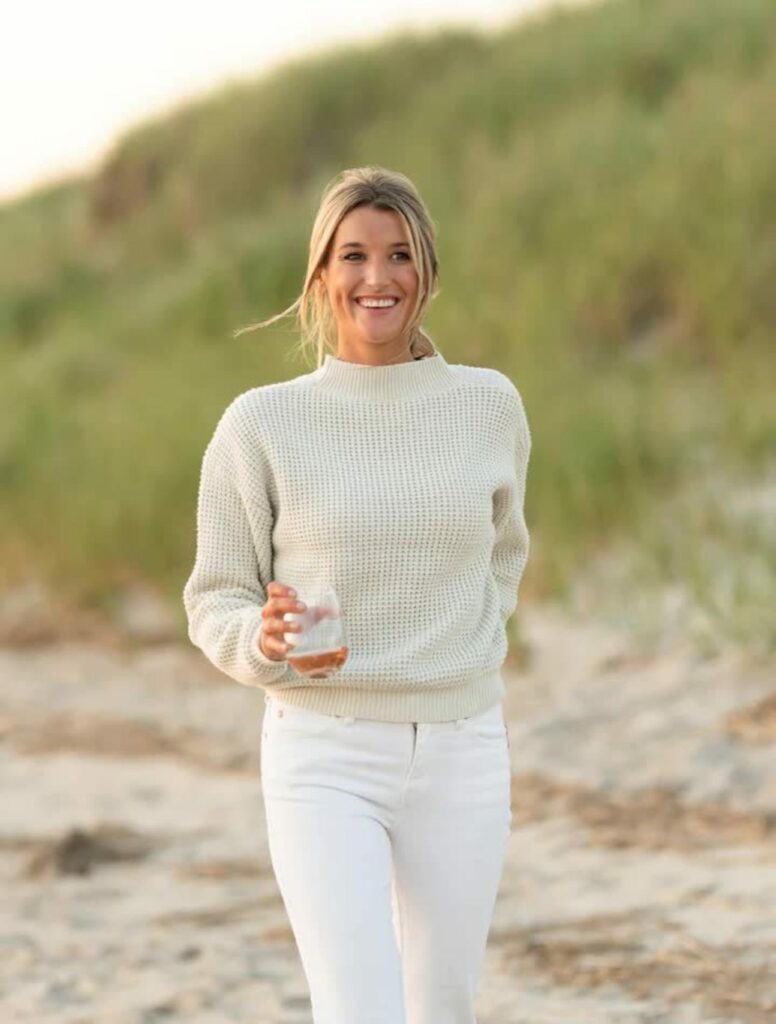 A blonde woman on a beach wears a cream-colored, waffle-knit, mock-neck sweater.