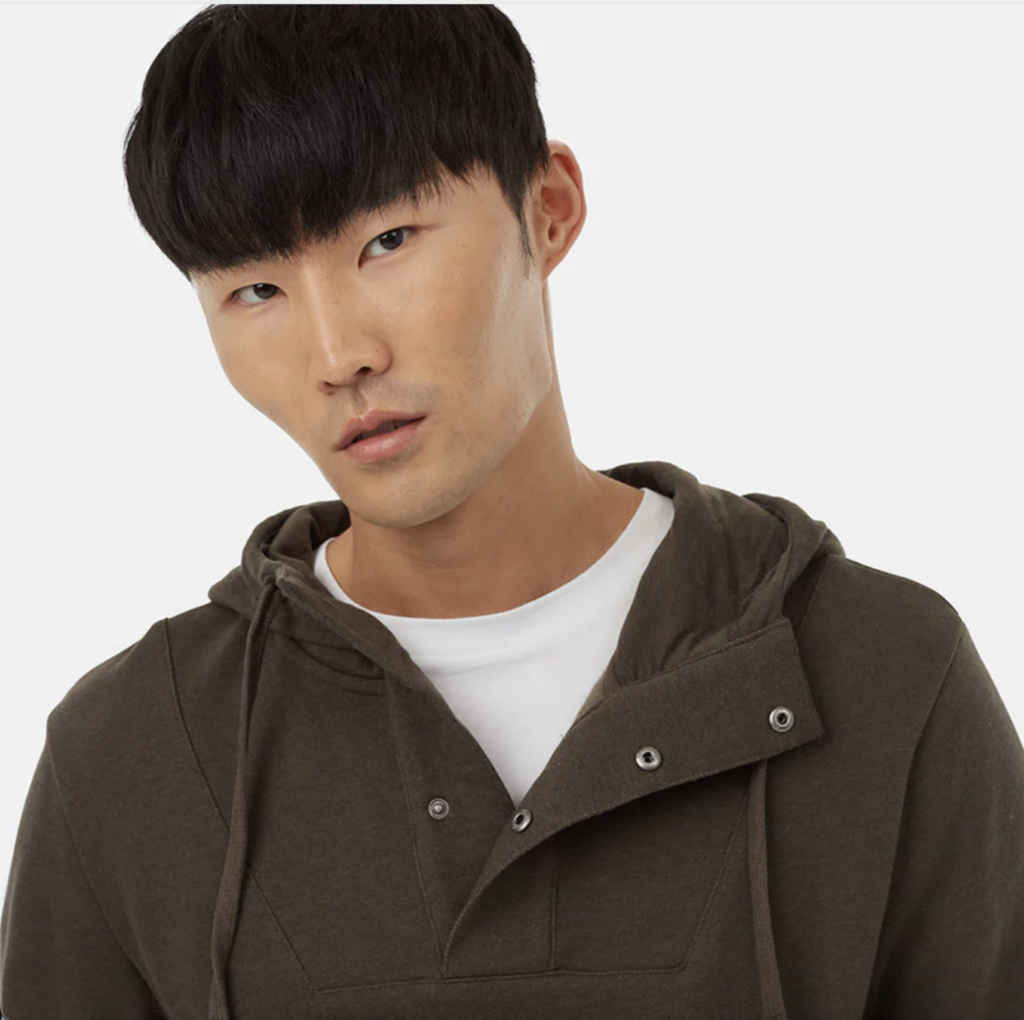 A young man with dark hair and a strong jaw wears a hooded brown sweatshirt with detailed snap closures.