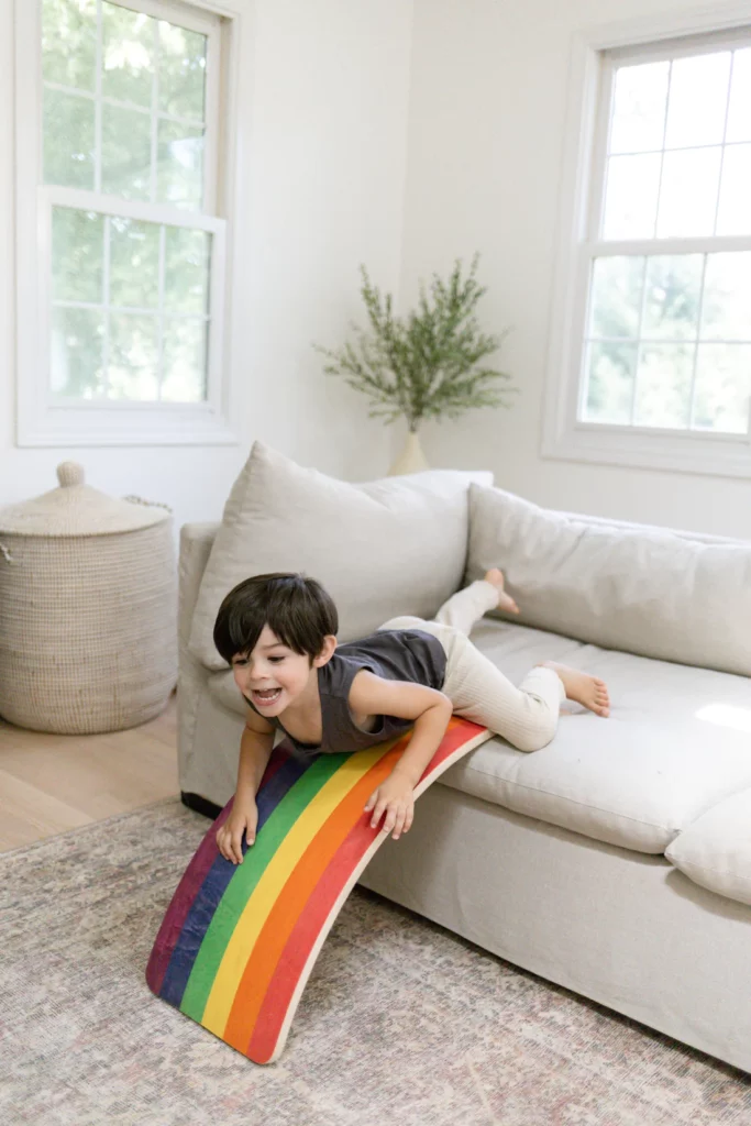 A smiling child is about to slide down a small wooden board, painted like a rainbow, that he has positioned off the sofa in a living room. 