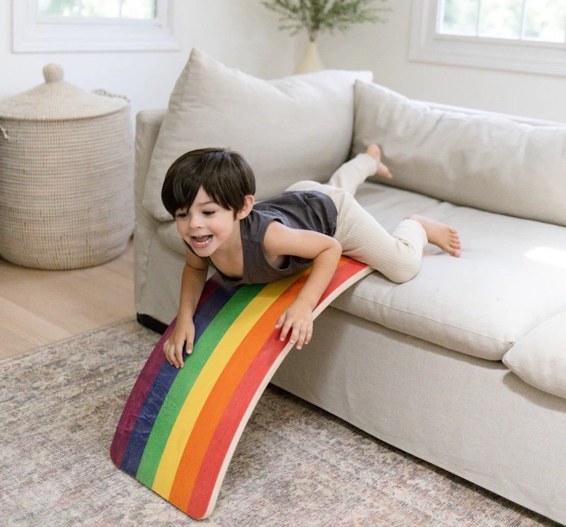 A smiling child is about to slide down a small wooden board, painted like a rainbow, that he has positioned off the sofa in a living room.