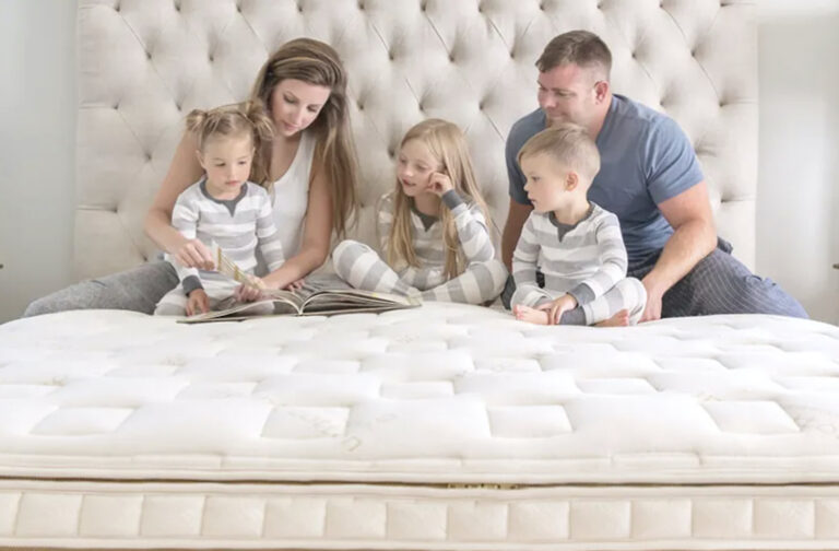 A woman, a man, and three small children, all blonde and wearing pajamas, sit on an organic mattress in front of a tufted cream-colored headboard. Image Courtesy Naturepedic