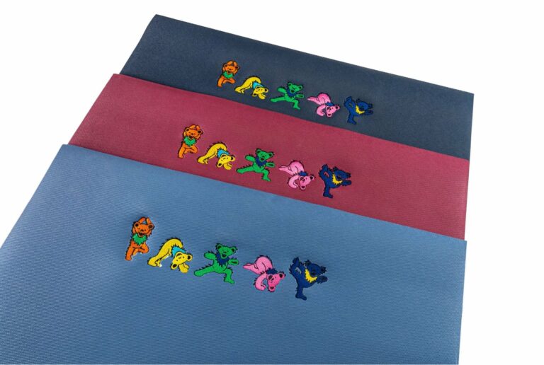 Three yoga mats are lined up together, one blue, one deep red, and one navy, on a white background. All have five bears from the Grateful Dead bears doing yoga poses embroidered on the tops of the mats.