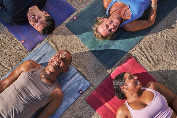 Four people ranging in age, gender, and skin tone lay smiling on their backs, faces up, on colorful rubber yoga mats. Their heads are close together and they wear athletic clothes.