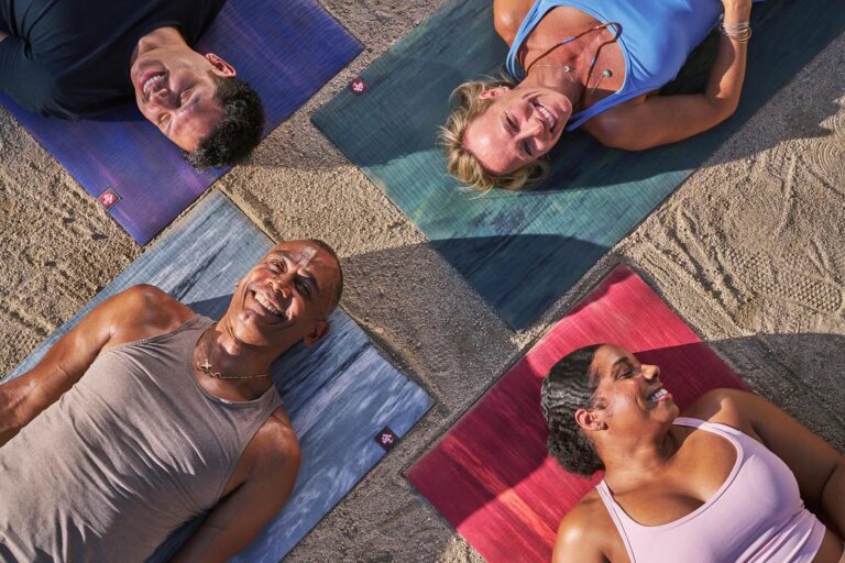 Four people ranging in age, gender, and skin tone lay smiling on their backs, faces up, on colorful rubber yoga mats. Their heads are close together and they wear athletic clothes.