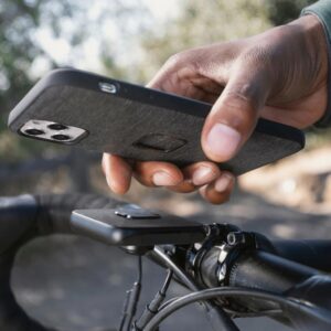 A person’s hand holds a cell phone in a gray case over a small phone mount connected to bicycle handlebars.