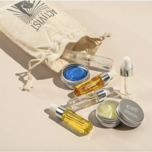 A small burlap bag with the word Activist on it is open on its side with small assorted vials and tins of skincare products.