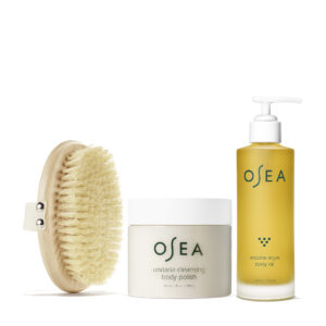 An oval-shaped wooden paddle brush with blonde bristles, a jar with a white cap and light brown contents labeled OSEA Undaria Cleaning Body Polish, and a tall jar with a white pump containing golden liquid labeled OSEA Undaria Algae Body Oil, all from sustainable seaweed skincare company OSEA.
