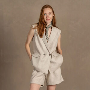 A red-headed woman shown from the knees up wears a matching beige linen shorts and vest, with a green checkered linen kerchief around her neck, and smiles at the camera. She stands in front of a brown photo background and wears linen clothes made to order from Lithuanian brand Menique.