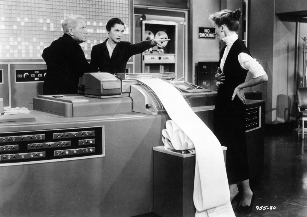 Spencer Tracy and Katharine Hepburn in the 1957 movie, Desk Set. They appear to be arguing over a giant computer with lots of paper printing from it.