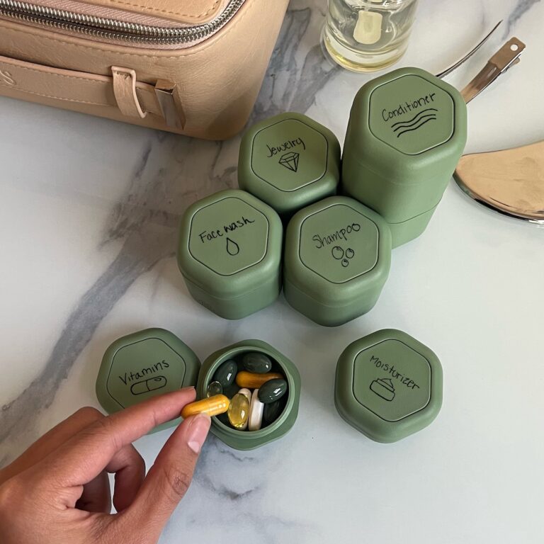 A white marble counter has seven opaque green hexagonal capsules on it labeled with the names of different products: Face Wash, Jewelry, Shampoo, Conditioner, and Moisturizer. Another capsule is ope and reveals vitamins and supplements inside, and a personโs hand is shown removing a vitamin pill.