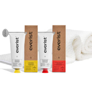 Two white tubes shown next to two cardboard boxes with a white towel in the background. The white tubes ach have a metal key at the top to help get all the product out of the tube. The products have the brand name Everist. The tube with the yellow top says “The Shampoo Concentrate” and the tube with the red top reads “The Conditioner Concentrate.”