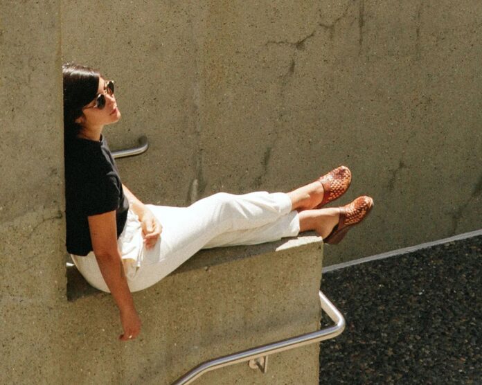 A woman in a black cotton tee shirt and white pants sits on a parapet above a staircase, facing forward.