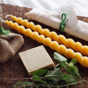 Two long and twisty beeswax candles, a small square beige soap, a small burlap package tied with green string, and a small wrapped packaged with a card reading Mile End Soap and Candle Supply are displayed alongside fresh rosemary and mint on a marble countertop.