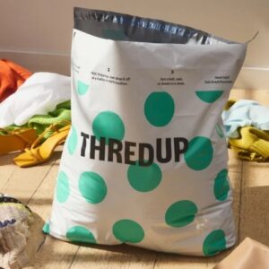 A white plastic bag with turquoise polka dots and the word THREDUP on it in the middle of a wooden floor, surrounded by women’s clothes and shoes.