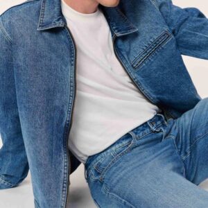 A person reclines on the ground, wearing matching tight bluejeans and a zip-up jean jacket and a white tee shirt.