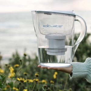 A person's hand holding a water pitcher branded Epic Water Filters in front of a patch of black and yellow flowers.
