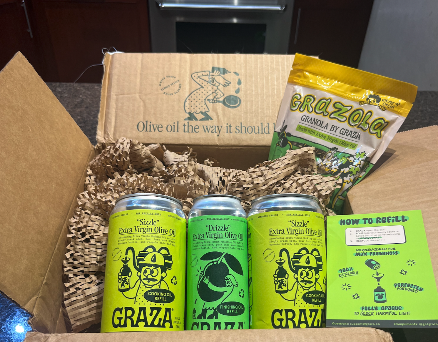 An open cardboard box on a kitchen counter. The box contains three green refill cans of Graza Extra Virgin Olive Oil, an instruction leaflet on how to refill your cans, and a packet of Graza Granola.