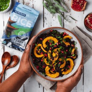 A person’s hands cradle a bowl of cooked dark-purple rice with sliced rings of squash, pomegranate seeds, and green onions. In the background, a back of Lotus Foods Heirloom Forbidden Rice.