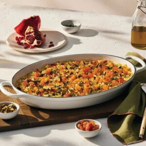 A white oval baking dish with a squash casserole inside, with garnishes on the side, including a pomegranate.