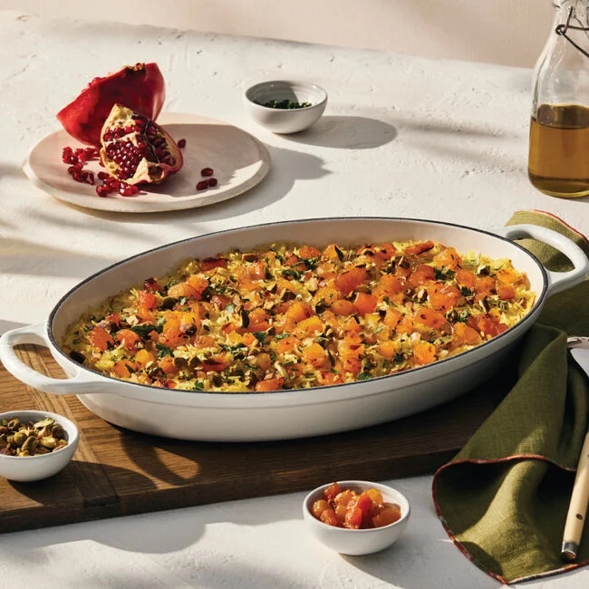 A white oval baking dish with a squash casserole inside, with garnishes on the side, including a pomegranate.