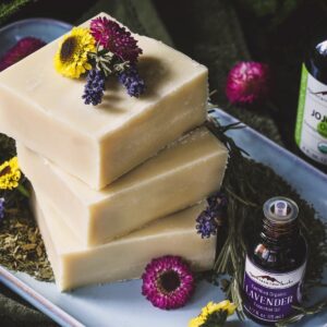 Three bars of soap on a plate, surrounded by bottles of essential oils from the brand Mountain Rose Herbs and colorful fresh flowers.