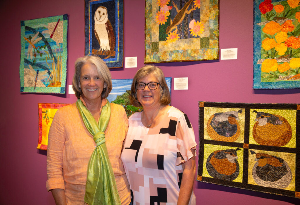 two ladies stand in front of a collection of quilts