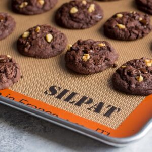 A baking sheet lined with a beige mat with a rust-colored rim that says SILPAT, topped with baked chocolate cookies.
