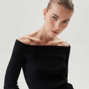 A woman with slicked back blonde hair and blue eyes wears a ribbed, off-the-shoulder black top.