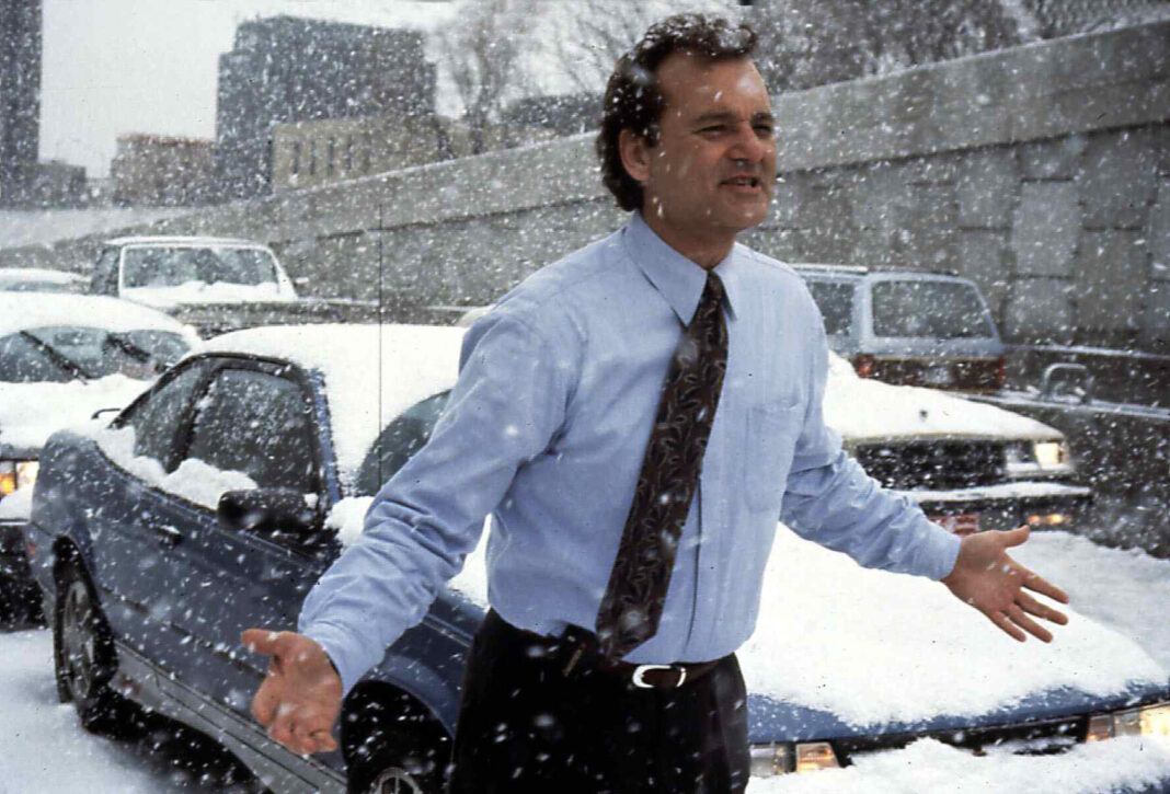 a man in a shirt and tie stands outside on a street full of parked cars in a heavy snowstorm