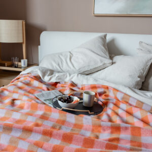 A bed with an orange pink and brown checked wool blanket on top, a newspaper, and a tray with a bowl of cherries and a mug on it.