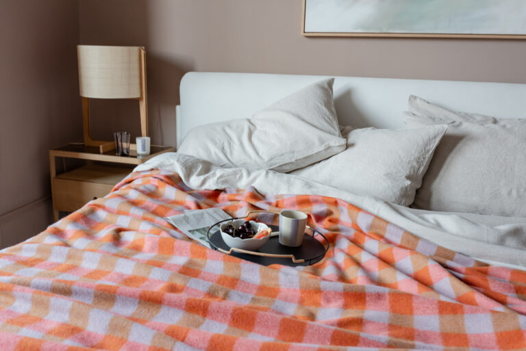 A bed with an orange pink and brown checked wool blanket on top, a newspaper, and a tray with a bowl of cherries and a mug on it.