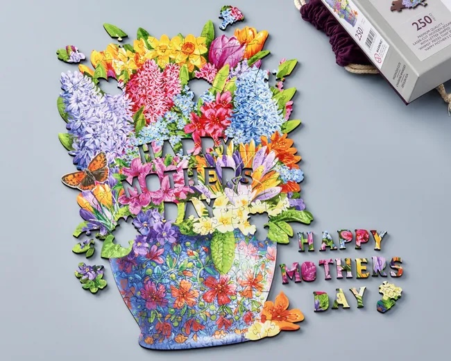 A mostly completed puzzle of a colorful bouquet of flowers. Puzzle pieces not yet placed in the puzzle are shaped into the letters that spell out “Happy Mothers Day.”