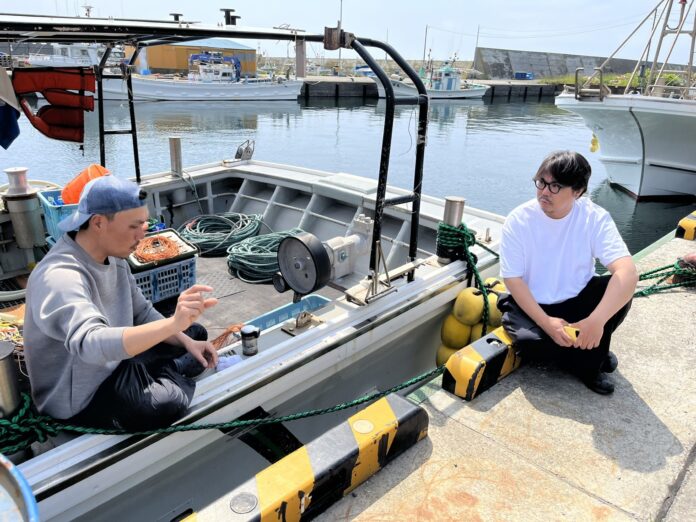 Shinji Murakami (right) sits on a concrete dock and talks to a fisherman in a gray boat.
