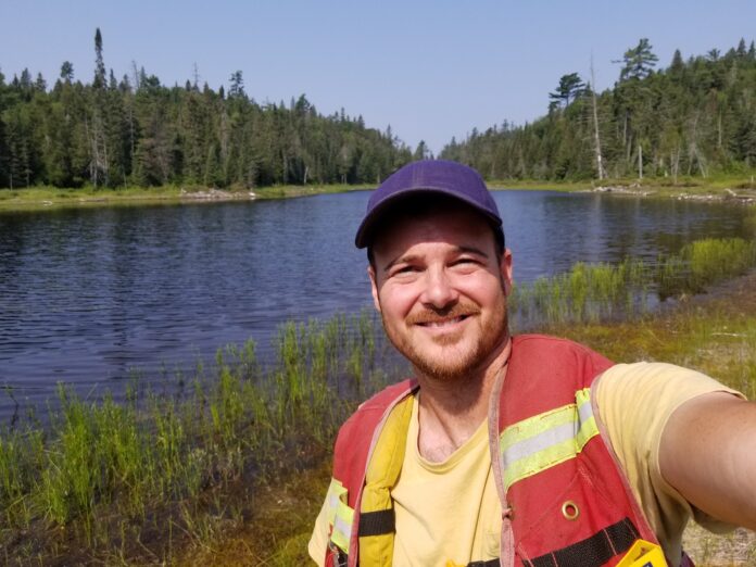Mark Balogh takes a selfie in front of a lake surrounded by trees.