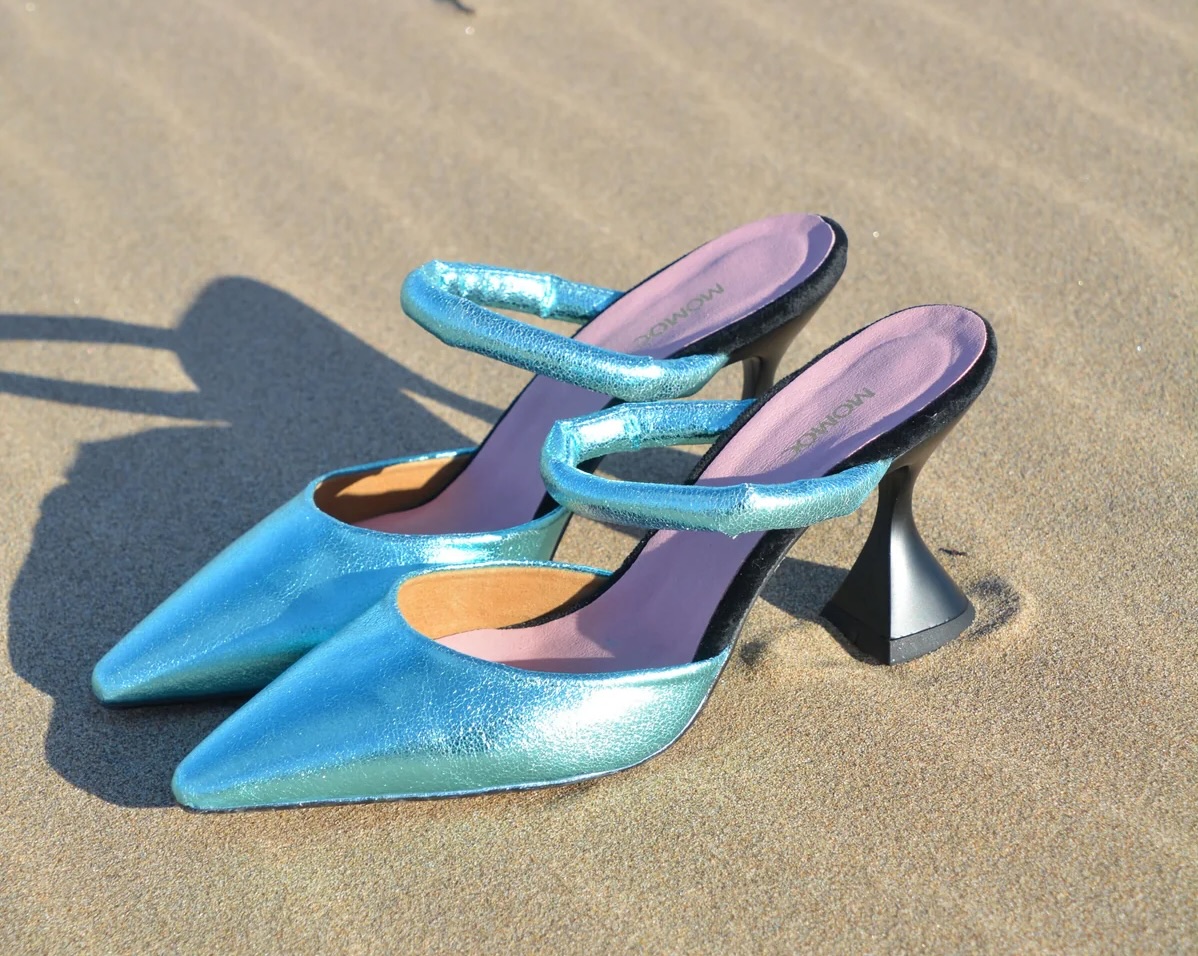 A pair of strappy metallic turquoise vegan leather high-heeled shoes on sand. The brand name MOMOC is written on the shoes’ insoles.