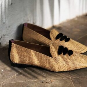 A pair of women’s raffia flat shoes with black laces in an outdoor corridor with whitewashed walls and palm frond shadows.