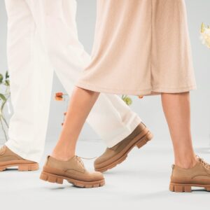 A man and a woman from the thighs down, wearing light colored clothing and walking towards each other wearing the same pair of beige lace-up shoes in a room filled with vases of flowers.