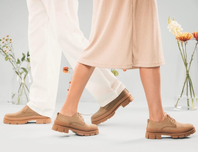 A man and a woman from the thighs down, wearing light colored clothing and walking towards each other wearing the same pair of beige lace-up shoes in a room filled with vases of flowers.