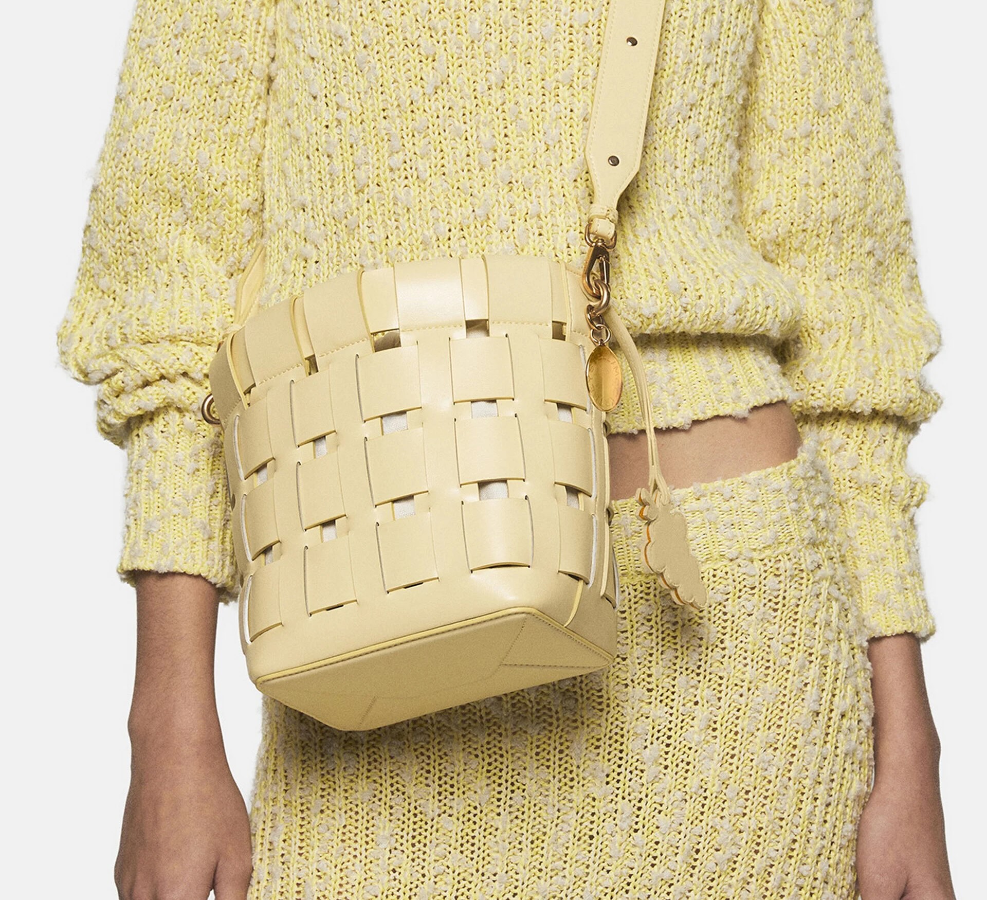 A person in a light yellow marled sweater and matching skirt holds a woven butter-colored leather bag with a keychain of grapes.