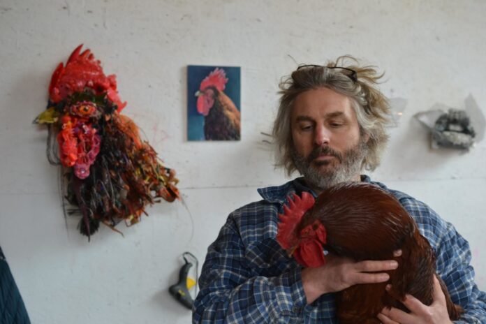 Thomas Deininger holds a roosted and sits in front of a painting and sculpture of a rooster