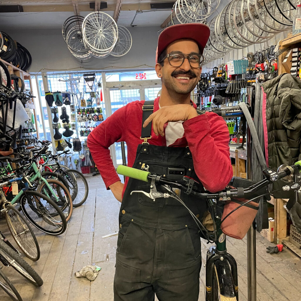 A man wearing a long-sleeve red shirt and black overalls poses in a bike repair shop.