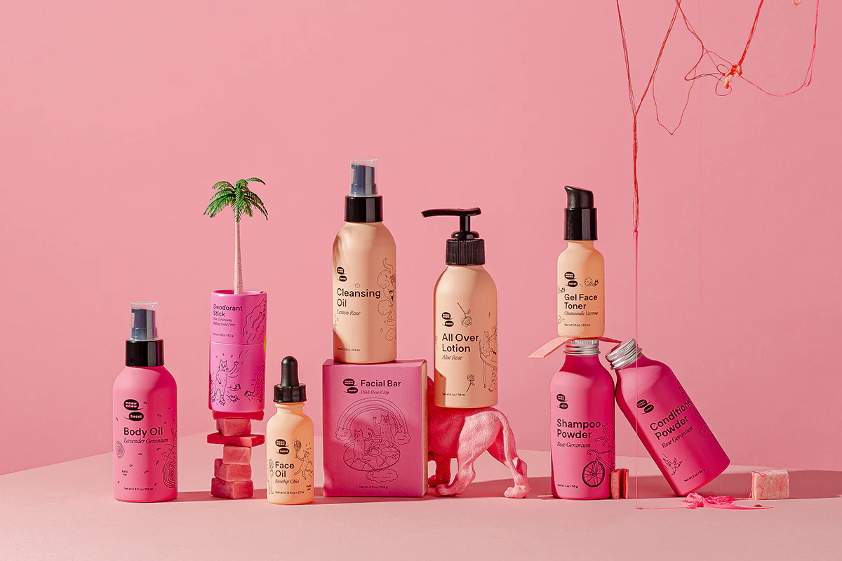 An assortment of Meow Meow Tweet brand products for face and body, all packaged in bright and light pink metal bottles and cardboard tubes.