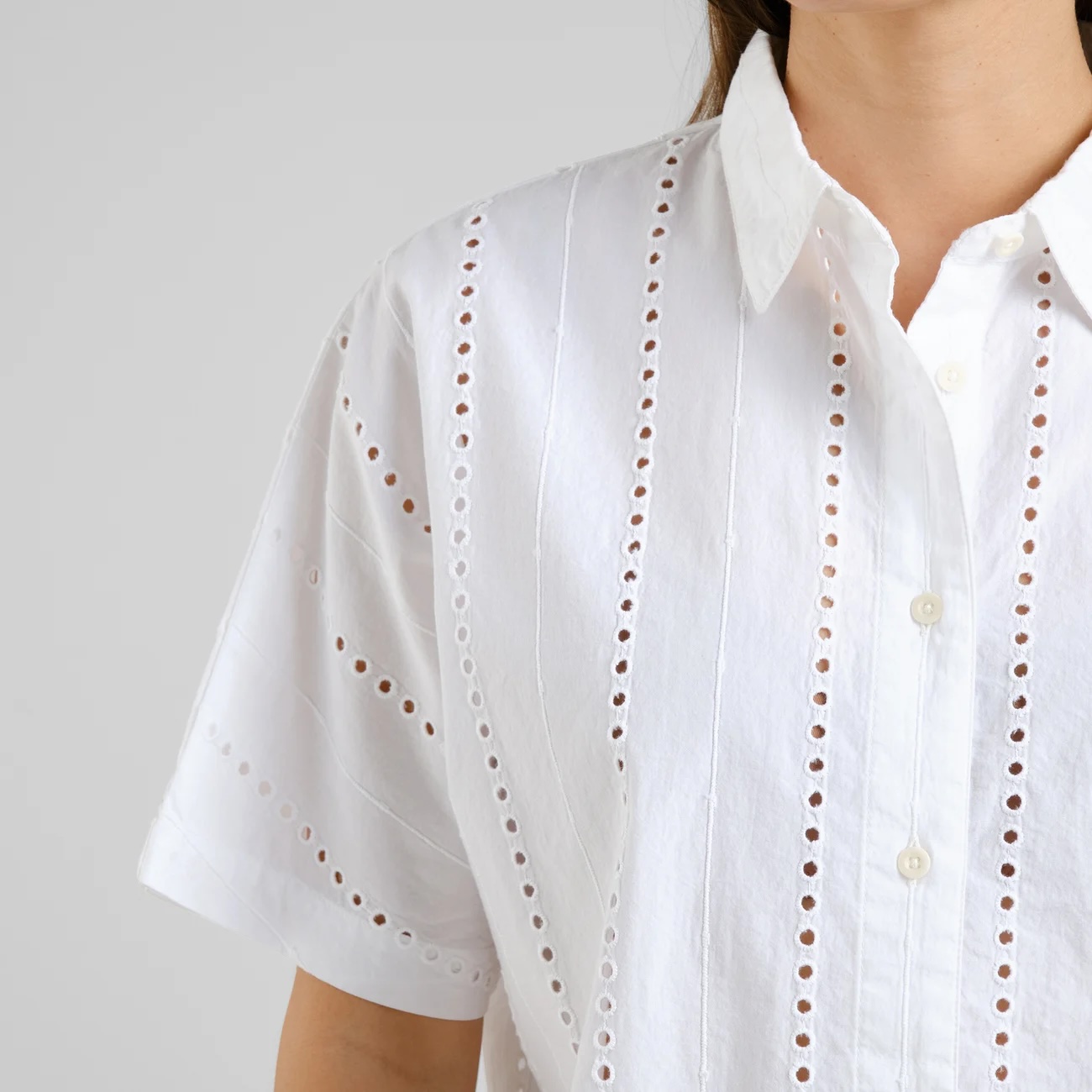 Close-up of a woman wearing a white short-sleeve button-up shirt featuring vertical rows of delicate eyelet embroidery and a pointed collar, set against a neutral grey background.