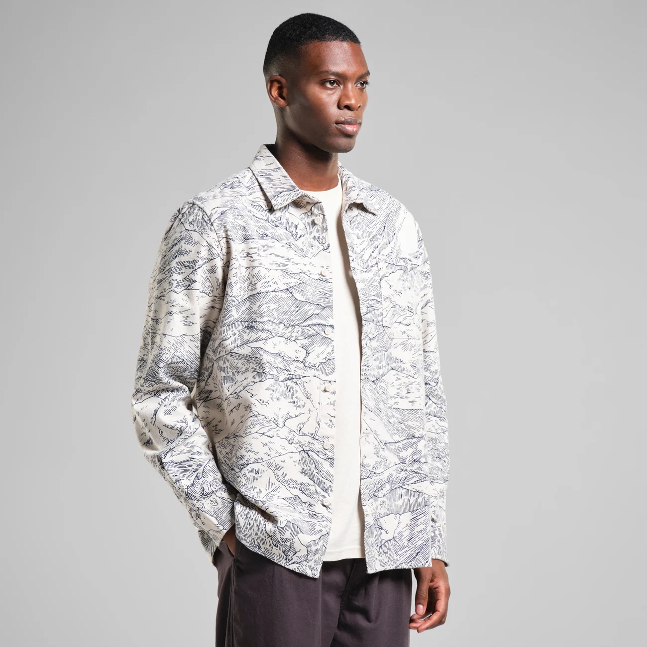 Man wearing a cream and navy printed jacket with an illustrated mountain landscape pattern, paired with a light beige t-shirt and dark trousers, standing against a neutral grey background.