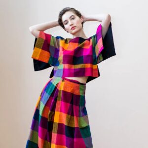 Woman wearing a vibrant, multicolored checkered two-piece set with wide-leg pants and a boxy top, posing confidently with hands behind her head against a neutral background.