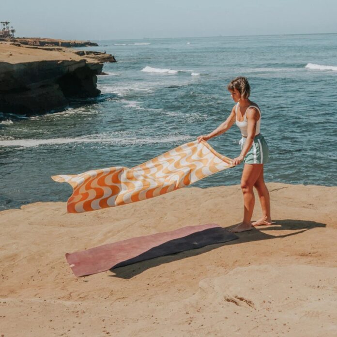 A woman unfurls an orange and white beach towel while looking out over the ocean