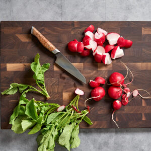 Fresh radishes, some whole and some chopped, with their leafy greens on a dark wood cutting board beside a chef’s knife with a burl wood handle and the brand name Steelport engraved on the blade.