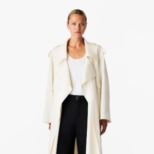 A blond woman wears an elegant trench coat in an off-white color over a white scoop neck top and black pants, made by Another Tomorrow Apparel.