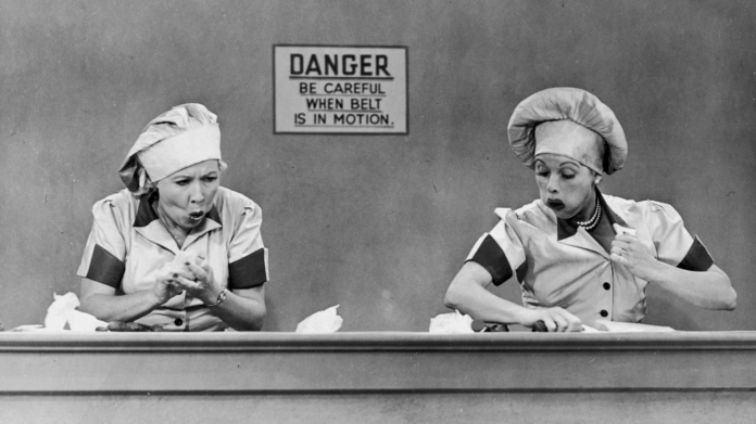 A black and white still image from the I Love Lucy tv show showing two women in uniforms standing in front of an assembly line.