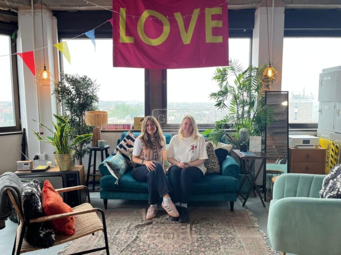 two women sit on a couch under a pink banner that reads "love"